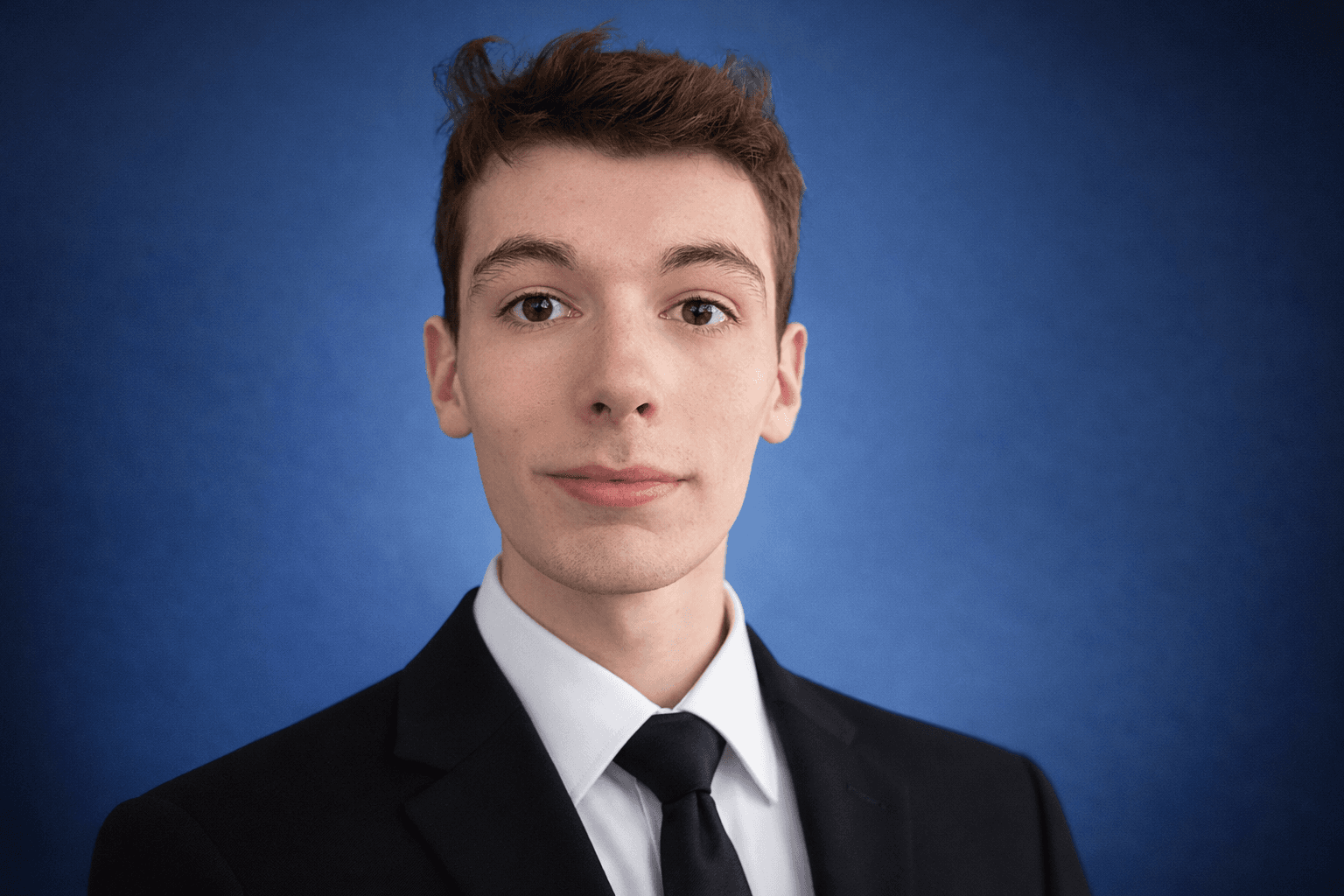 Professional headshot of a young man in a black suit against a blue background.