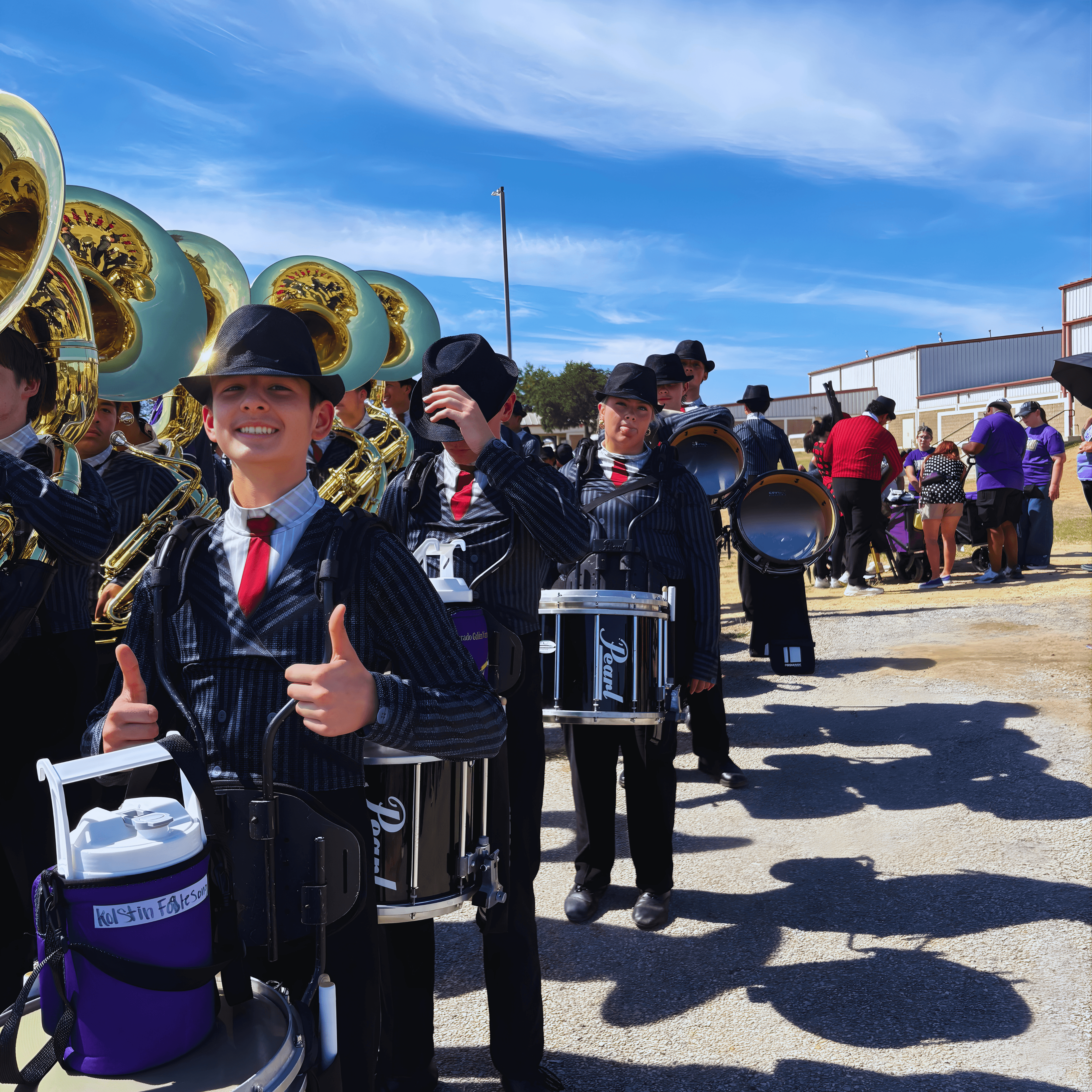 Smiling drummer in pinstripe suit and fedora gives thumbs-up, surrounded by band members.