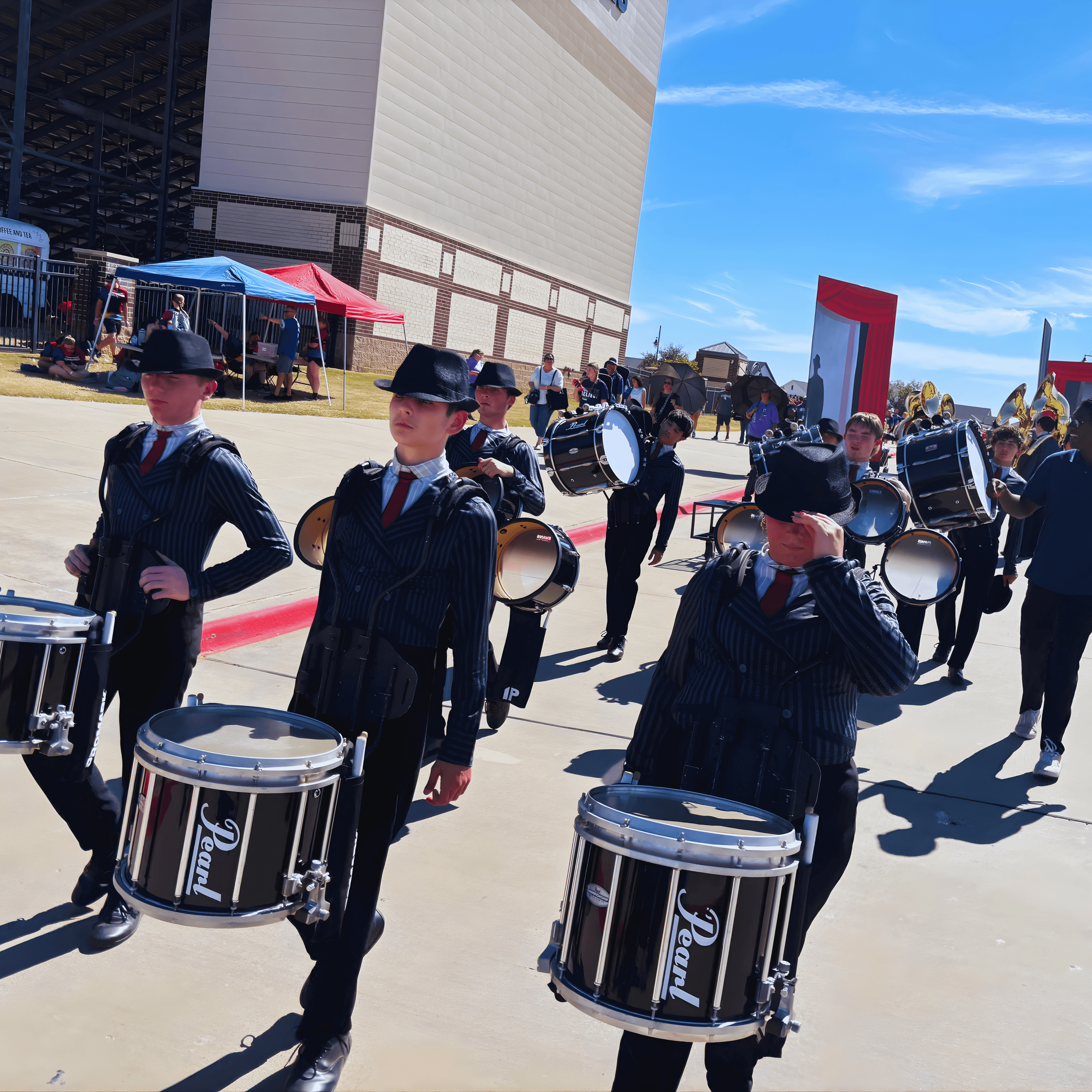 Marching band drumline in pinstriped uniforms and fedoras on a sunny day outdoors.